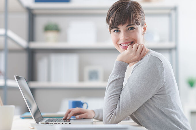Finanzbuchhalter (m/w/d) 61531086 - happy confident businesswoman sitting at office desk and working with a laptop, she is smiling at camera